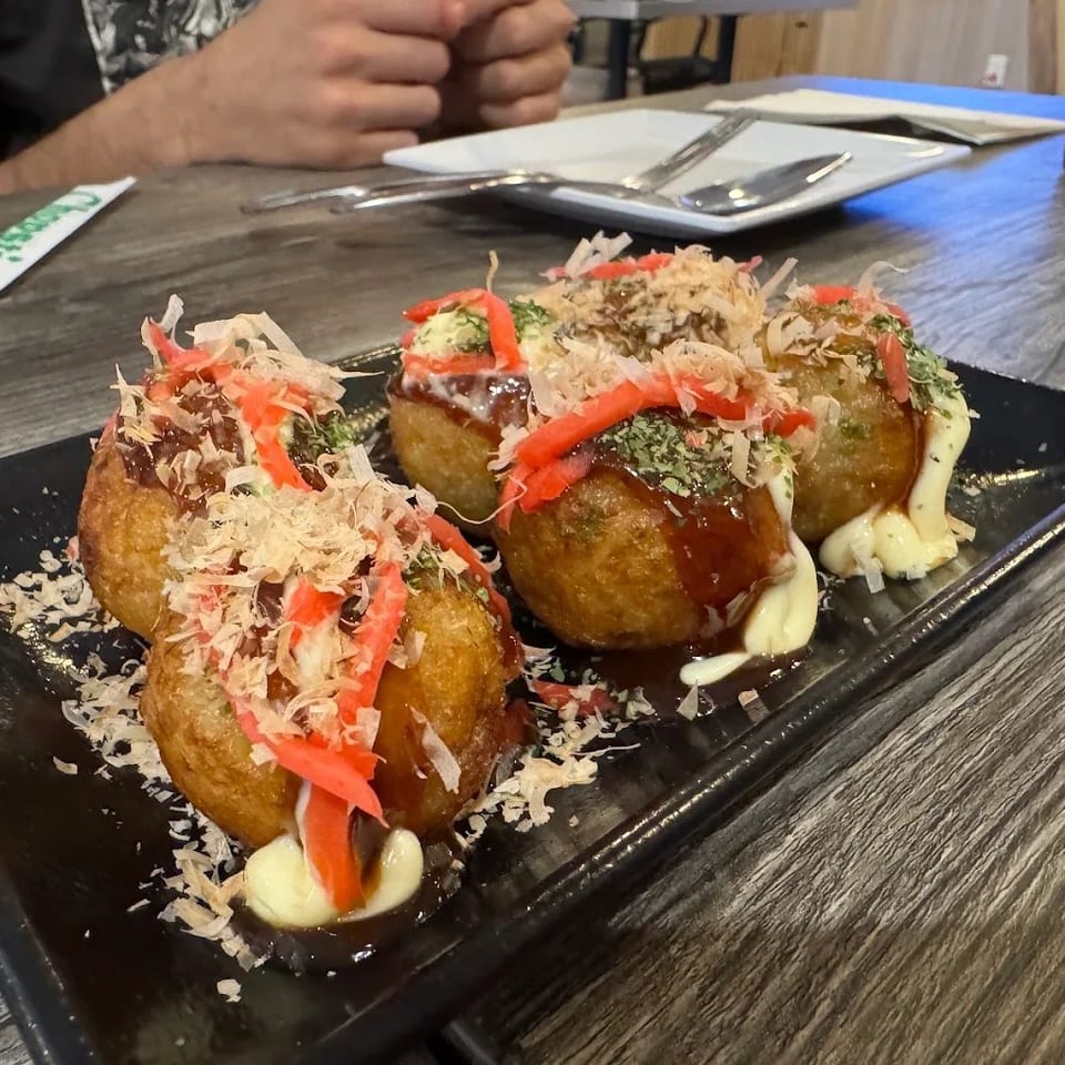 Three takoyaki balls topped with sauce, mayonnaise, bonito flakes, seaweed, and pickled ginger on a black plate.