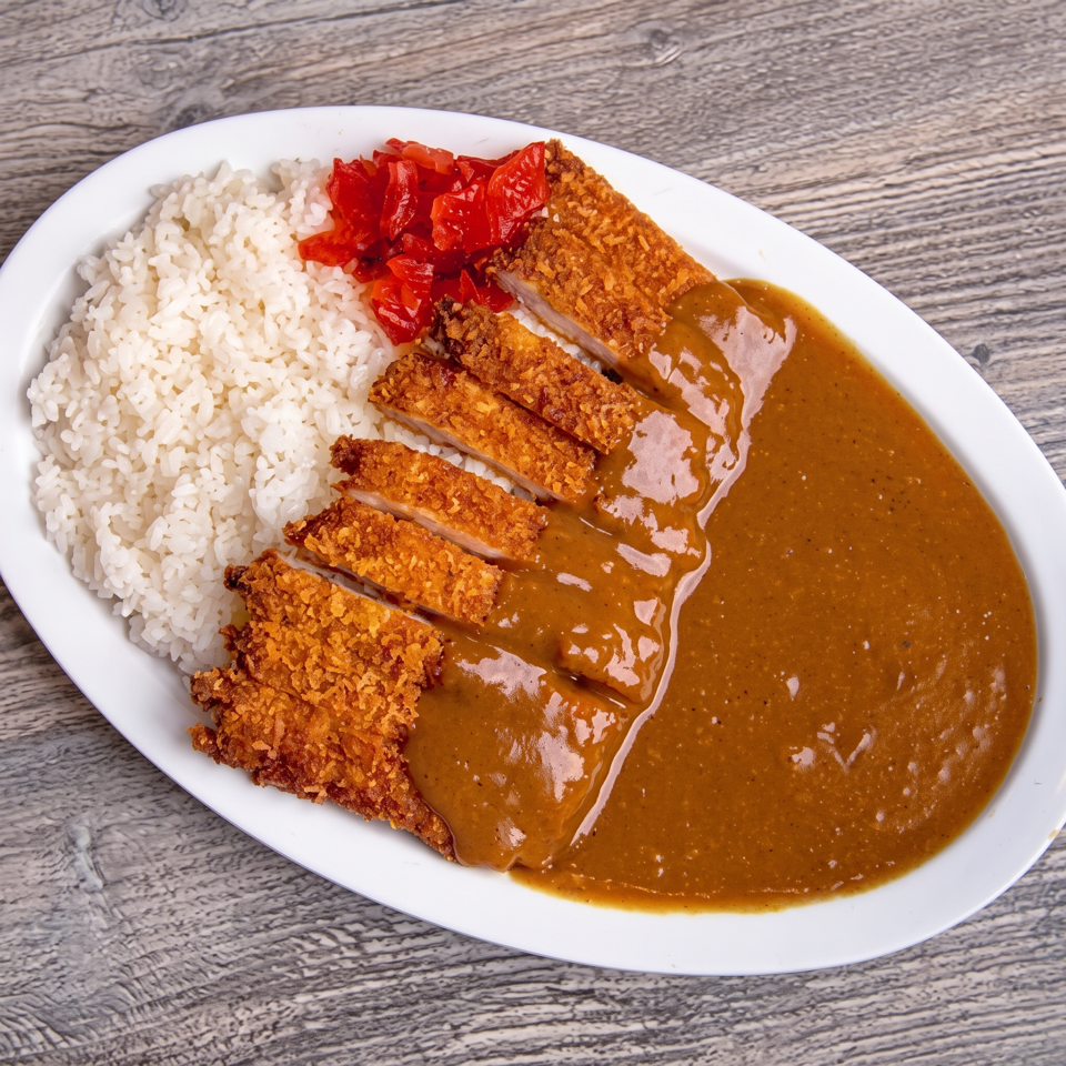 Plate of sliced crispy breaded cutlet with white rice, brown curry sauce, and red pickles on a wooden table.