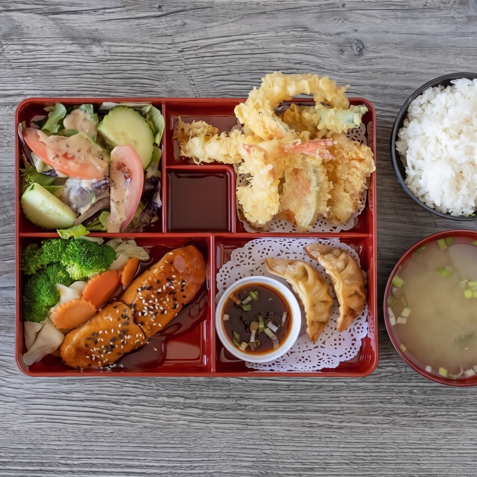 Japanese bento box with tempura, teriyaki chicken, salad, vegetables, dumplings, dipping sauce, rice, and miso soup.