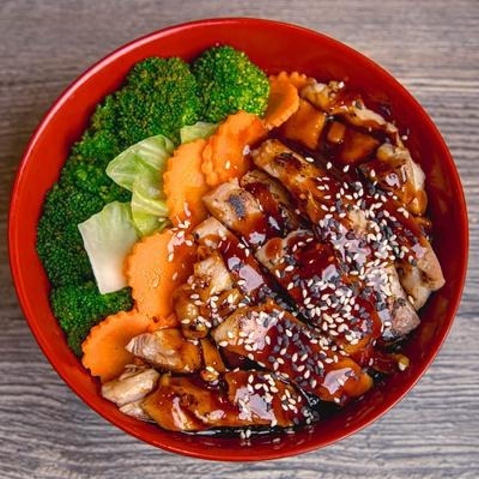 Red bowl of teriyaki chicken with broccoli, carrots, cabbage, and sesame seeds on a wooden table.